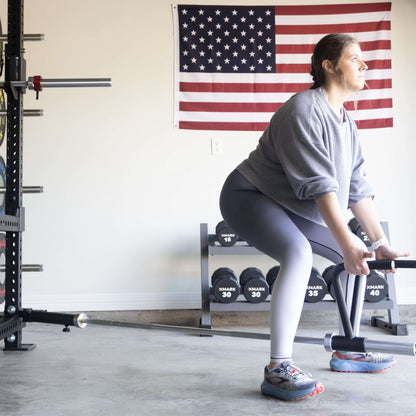 Woman using Viper landmine attachment for workout in garage, with American flag in background and dumbbells visible.