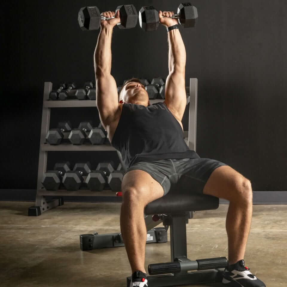 A man performing a shoulder press with dumbbells while seated on an adjustable weight bench, with weights in the background.