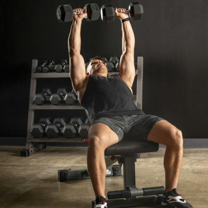 A man performing a shoulder press with dumbbells while seated on an adjustable weight bench, with weights in the background.